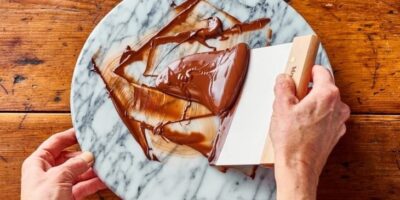 Hands spreading melted chocolate across a marble slab for tempering